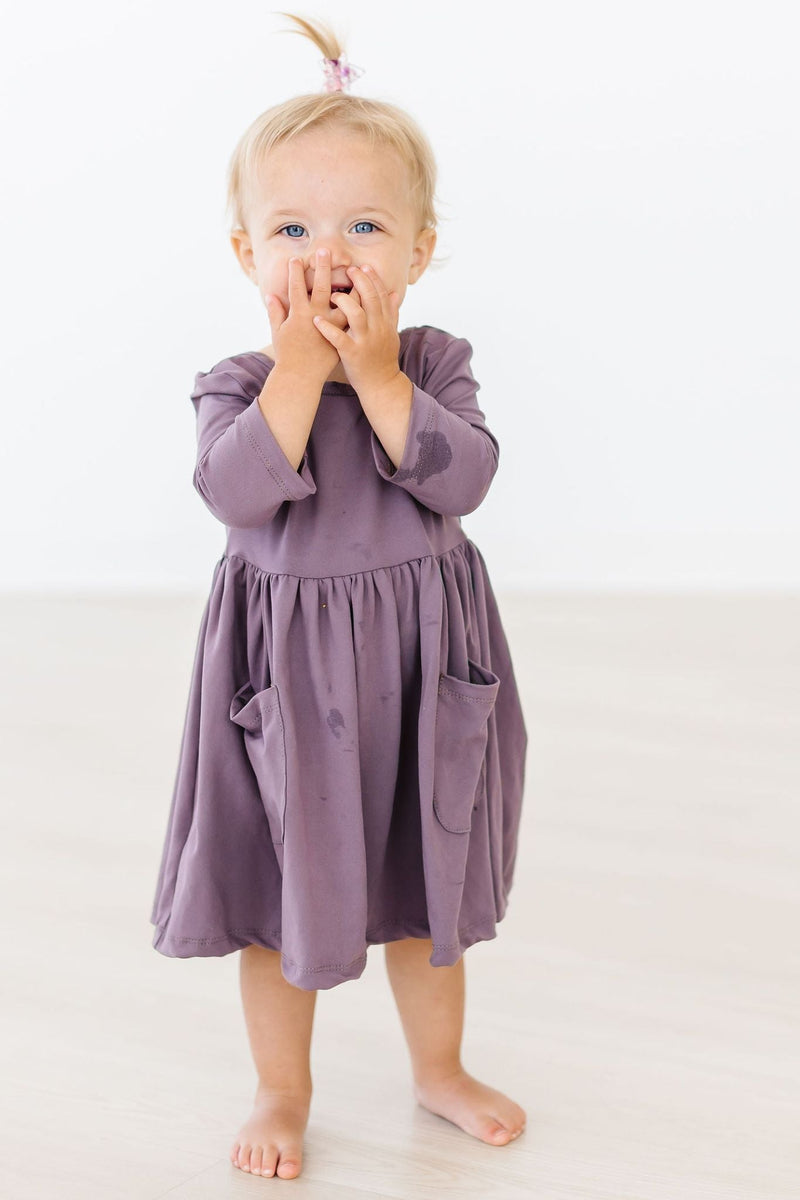 Toddler wearing a Vintage Violet 3/4 Sleeve Pocket Twirl Dress by Mila & Rose, playfully covering her mouth in a bright, clean space.