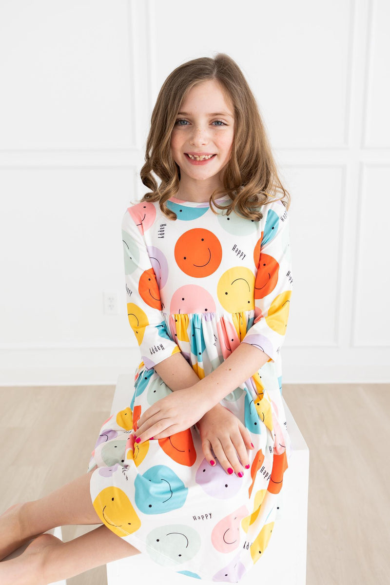 A young girl happily seated in the 'Happy Days' 3/4 Pocket Fun Twirl Dress for Girls, emphasizing the twirl-friendly and colorful design.