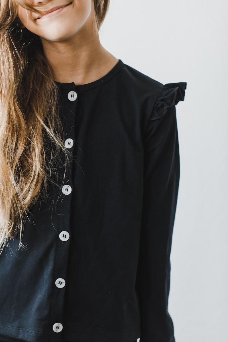 A child wearing a Black Ruffle Cardigan, featuring playful ruffles on the shoulder, styled with buttons down the front and a cheerful smile, perfect for layering in cooler weather.