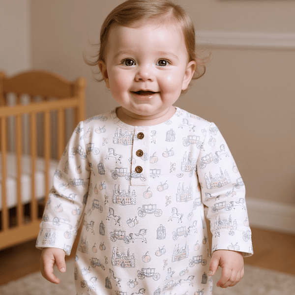 Smiling baby in white Fairy Godmother Ruffle Gown featuring carriage, castle, and pumpkin print with front snap placket.
