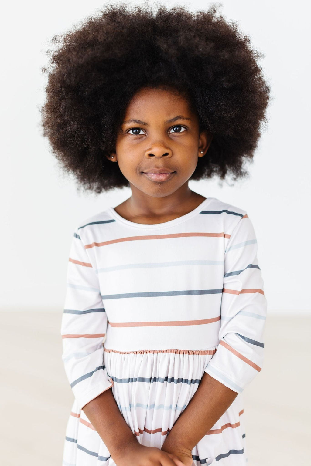 A young girl wearing the Fall Stripes 3/4 Sleeve Pocket Twirl Dress, featuring a variety of horizontal stripes. She has a beautiful curly hairstyle and is posing confidently, capturing the playful spirit of the dress.