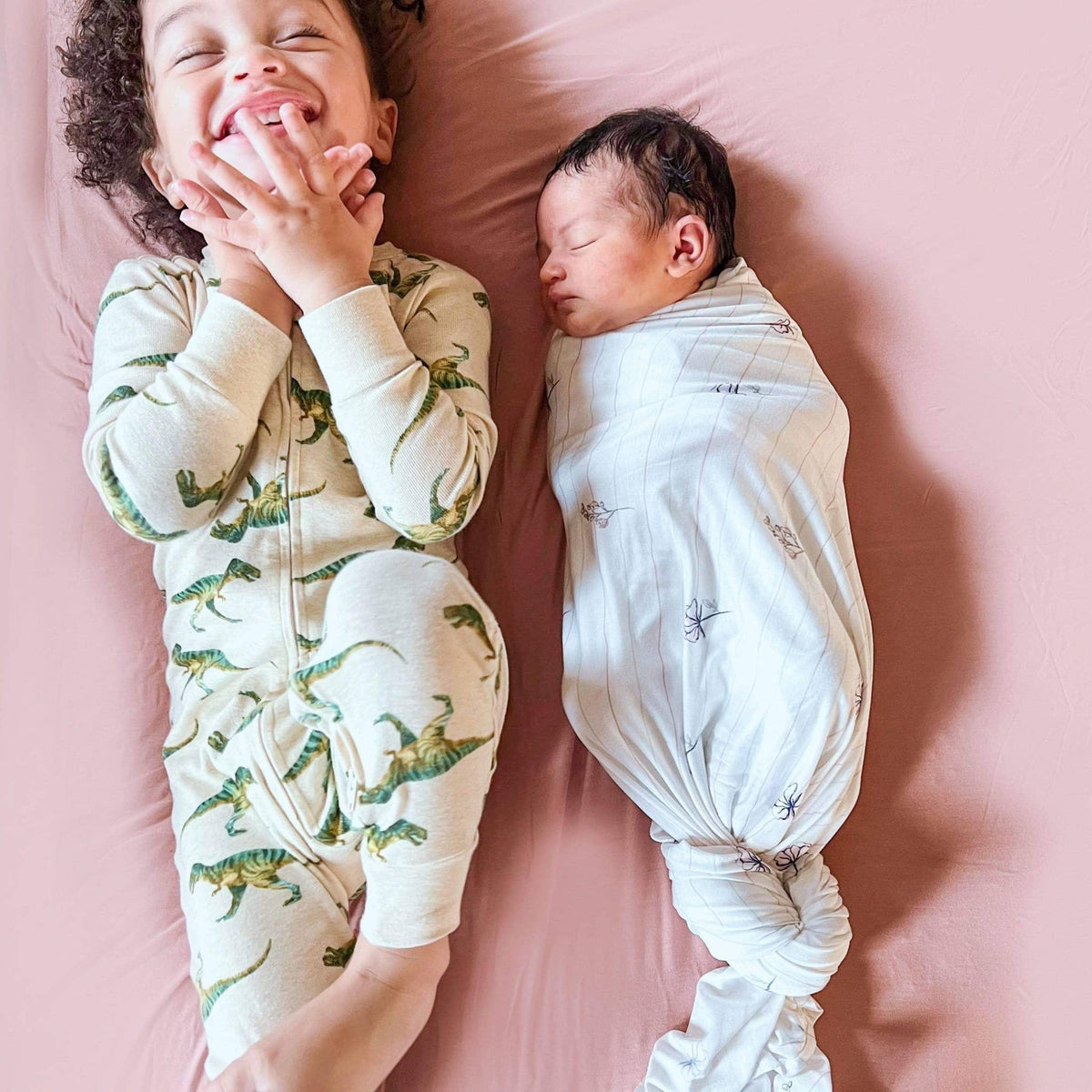 Newborn baby swaddled sleeping in flower pink baby swaddle blankets next to smiling toddler in dinosaur pajamas