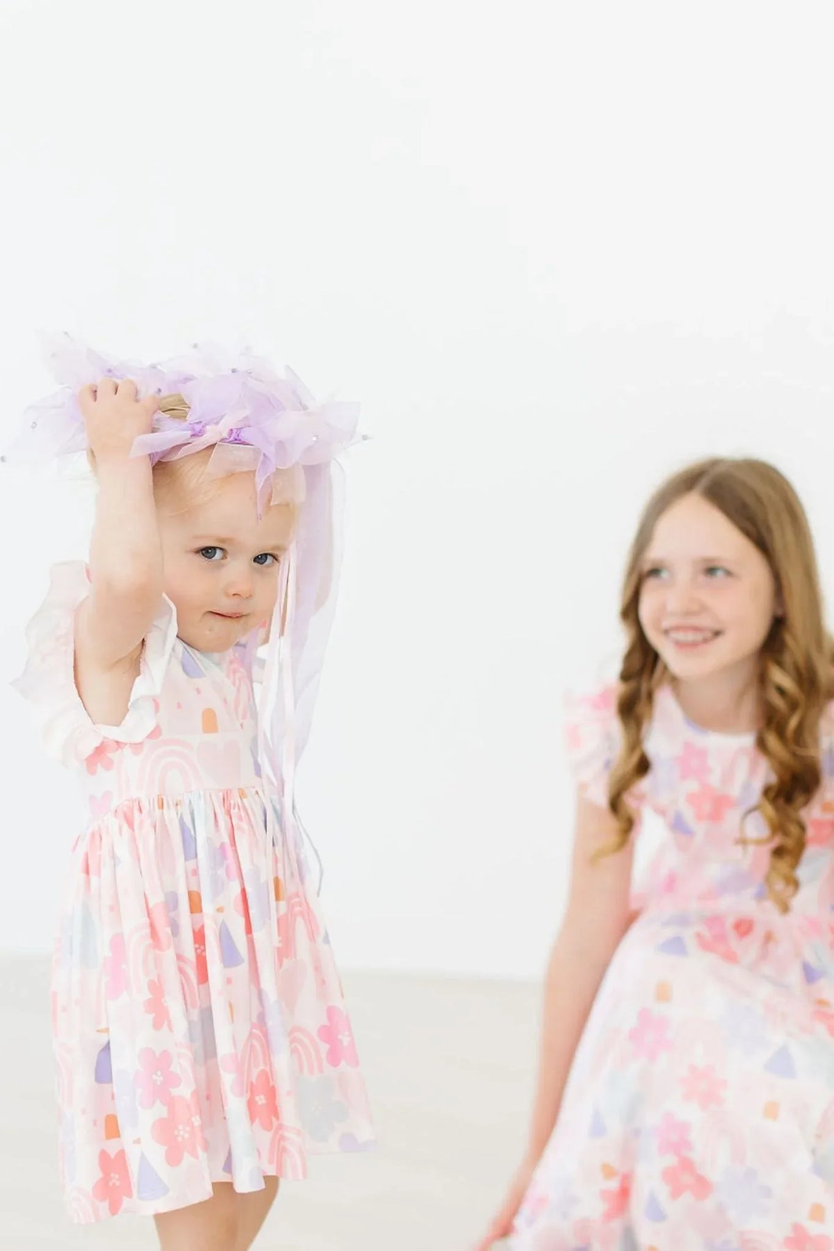 Toddler girl wearing a pastel floral flutter sleeve twirl dress for girls with a purple headband and older girl in matching dress