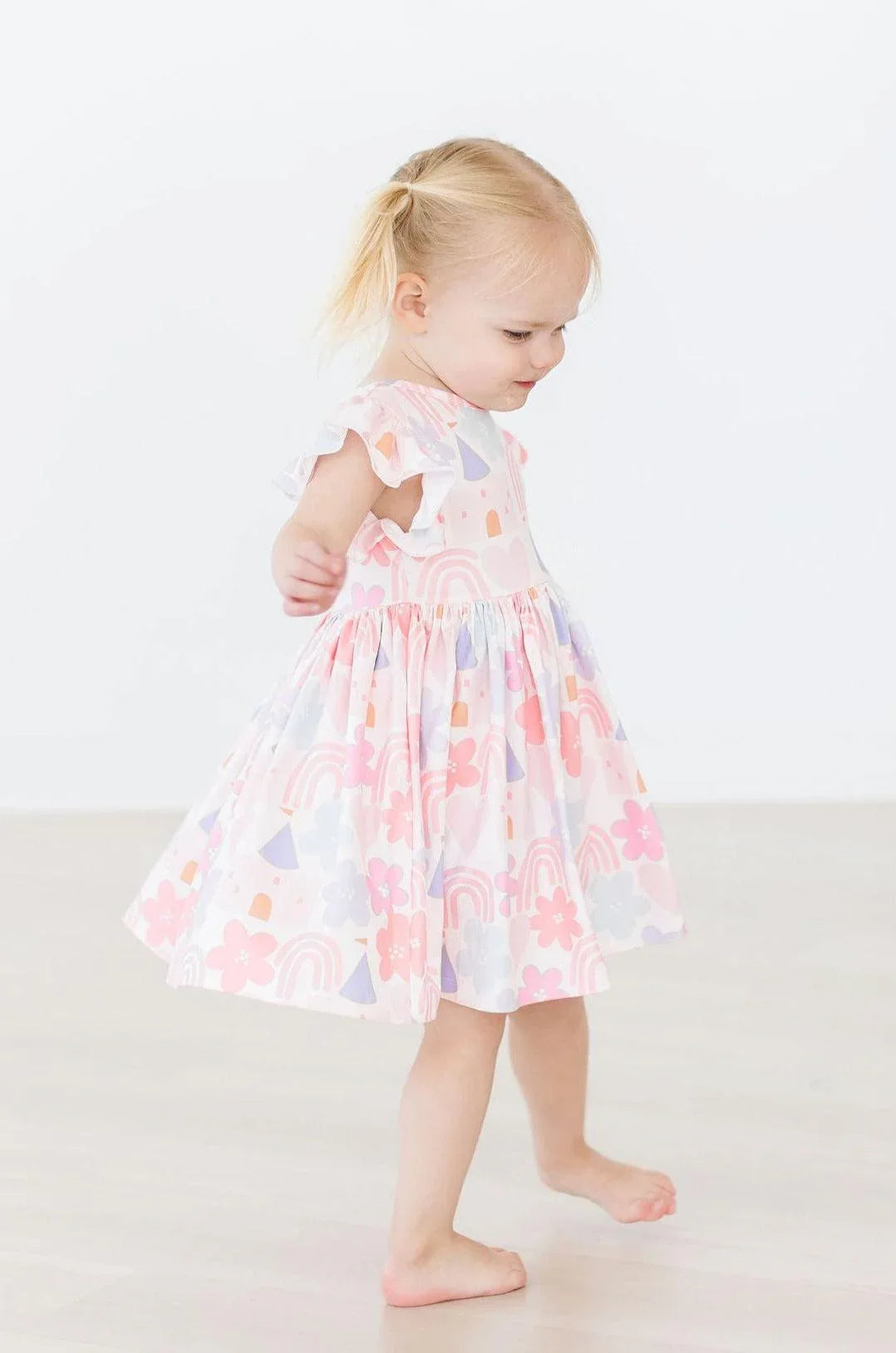 Toddler girl wearing Mila & Rose flutter sleeve twirl dress for girls in pastel pink with rainbow and flower print