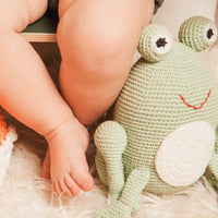Hand-crocheted green frog plush with big eyes and red smile sits on white rug beside a baby's legs. Soft organic cotton toy.