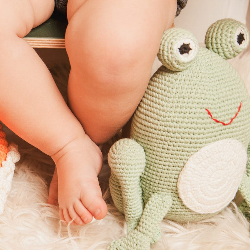 Hand-crocheted green frog plush with big eyes and red smile sits on white rug beside a baby's legs. Soft organic cotton toy.