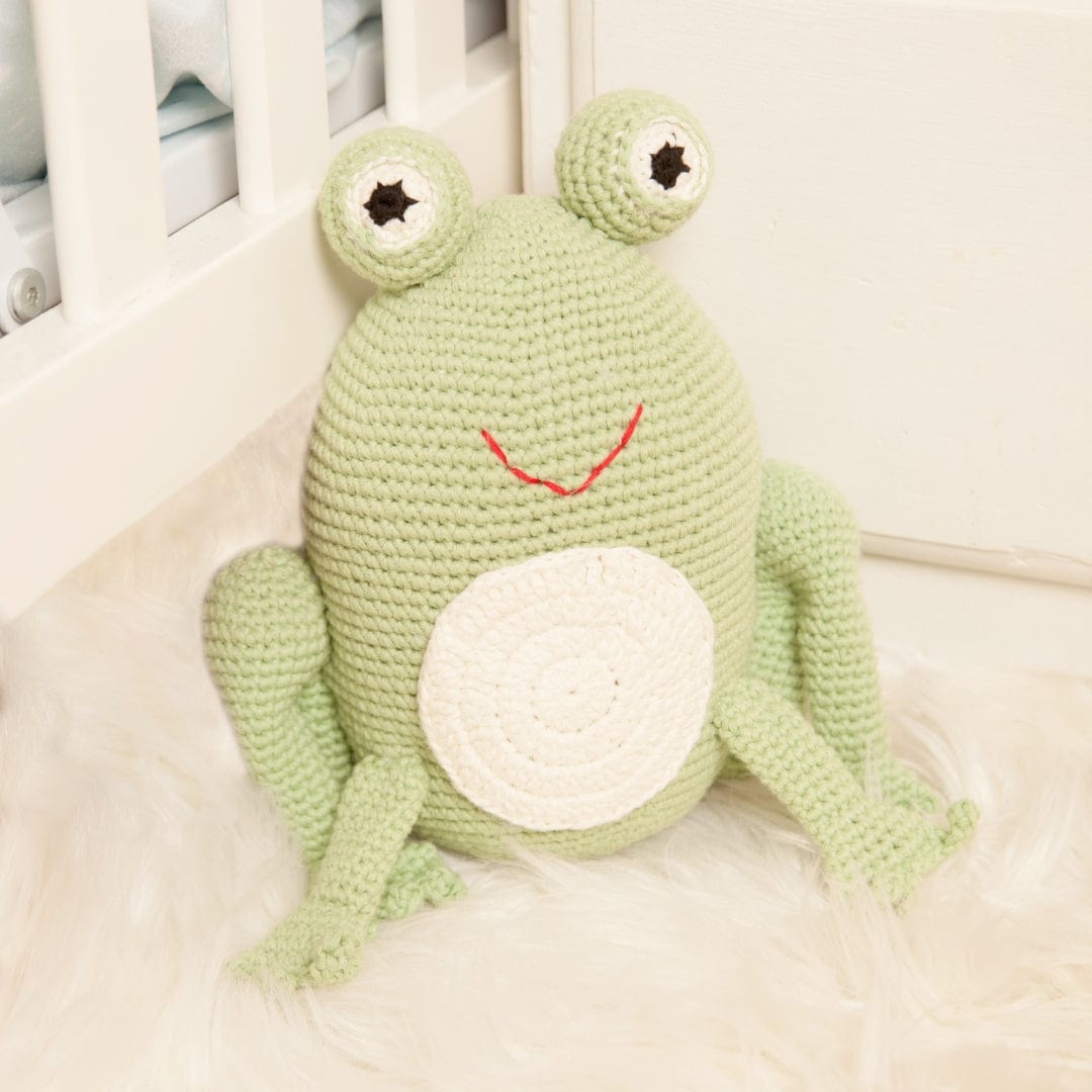 Soft green crochet frog with big eyes and red smile sitting on white fur rug. Handmade organic cotton toy for pretend play.