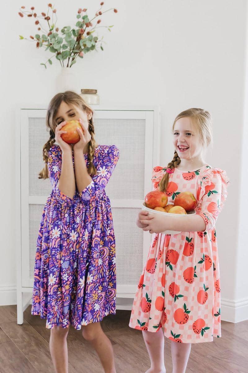 Two girls wearing purple and peach girls 3/4 sleeve twirl dresses holding and smelling apples indoors