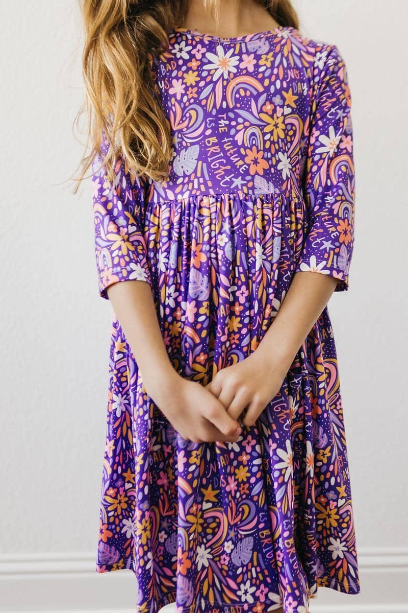 Girl wearing purple girls 3/4 sleeve twirl dress with floral and rainbow pattern standing with hands clasped