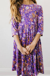 Girl wearing purple girls 3/4 sleeve twirl dress with floral and rainbow pattern standing with hands clasped