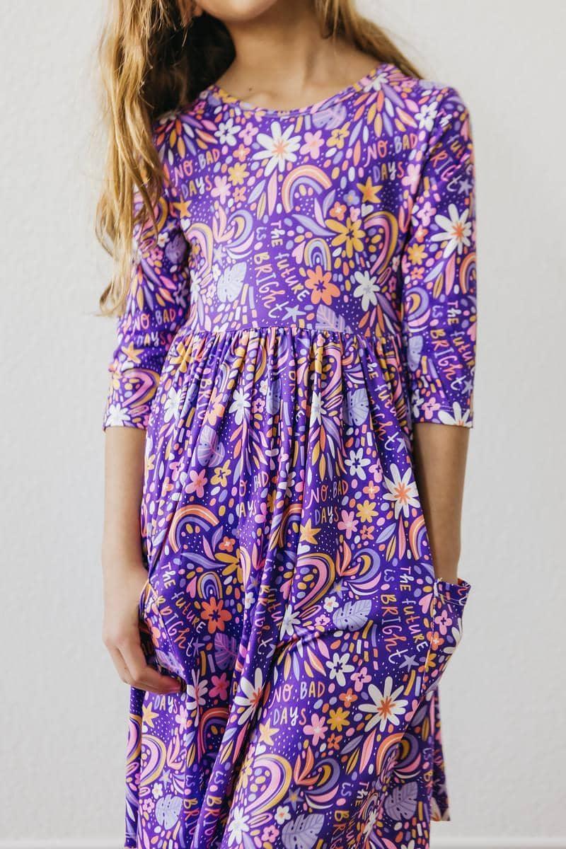Girl wearing purple girls 3/4 sleeve twirl dress with floral and graphic patterns holding dress pockets