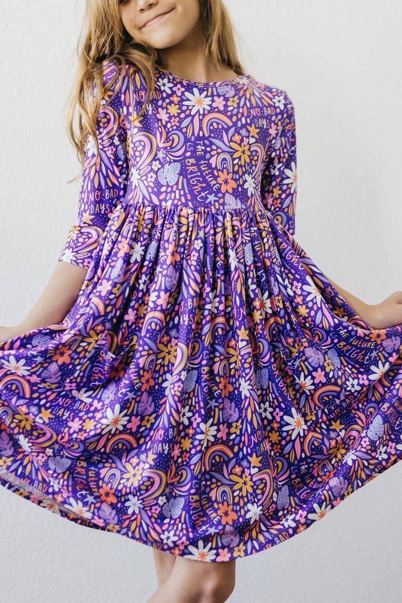Young girl wearing purple Mila Rose girls 3/4 sleeve twirl dress with floral and rainbow patterns holding dress out