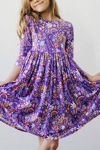 Young girl wearing purple Mila Rose girls 3/4 sleeve twirl dress with floral and rainbow patterns holding dress out
