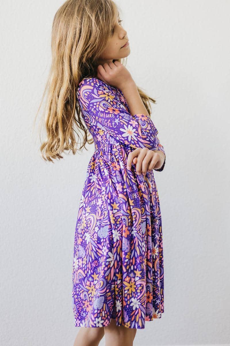 Young girl wearing purple girls 3/4 sleeve twirl dress with floral pattern standing sideways