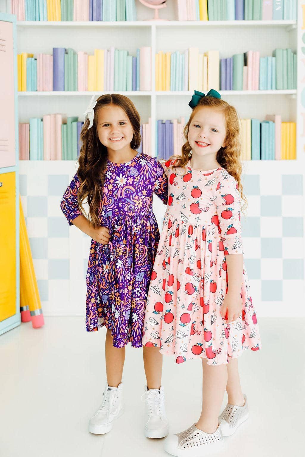 Two young girls wearing colorful girls 3/4 sleeve twirl dresses smiling and standing indoors
