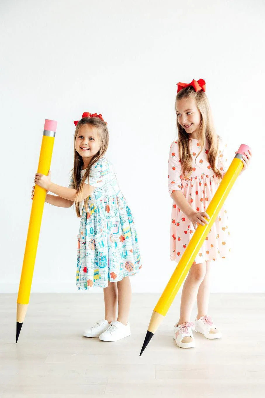 Two young girls wearing Mila and Rose girls apple print twirl dress in colorful and red apple patterns with sleeves holding large yellow pencils