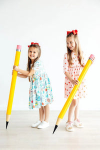 Two young girls wearing Mila and Rose girls apple print twirl dress in colorful and red apple patterns with sleeves holding large yellow pencils