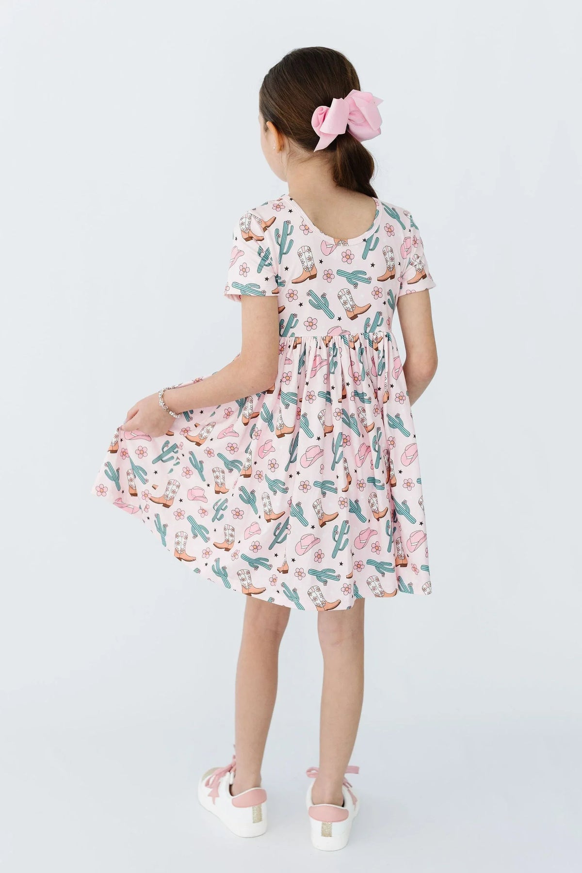 Young girl wearing pink girls cactus twirl dress holding hem and standing with back to camera