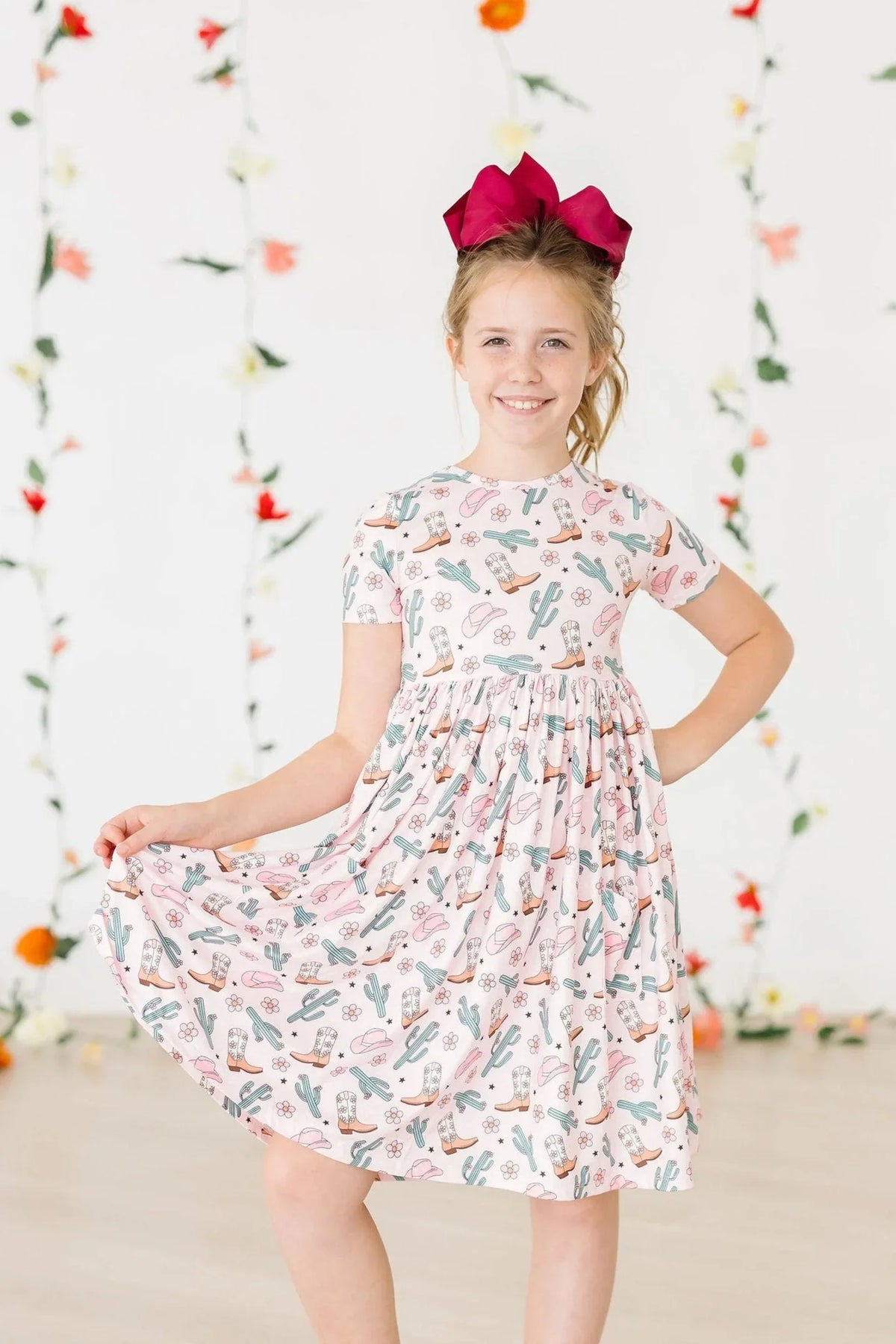 Young girl wearing a white girls cactus twirl dress with short sleeves and a pink and green cactus print smiling and holding the skirt
