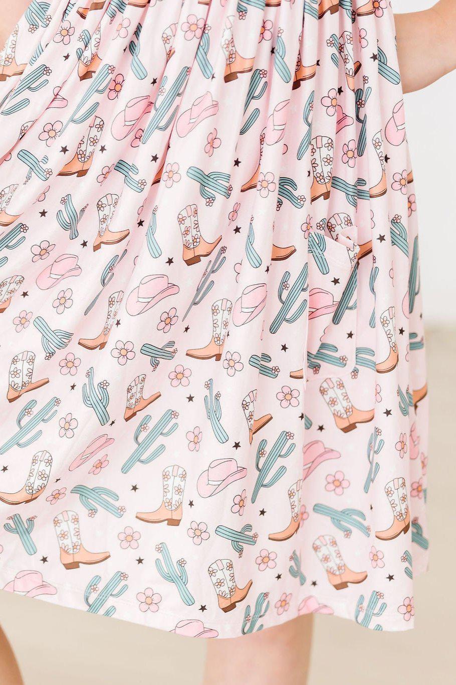 girl wearing pink girls cactus twirl dress with cowboy boot and cactus print holding dress sides