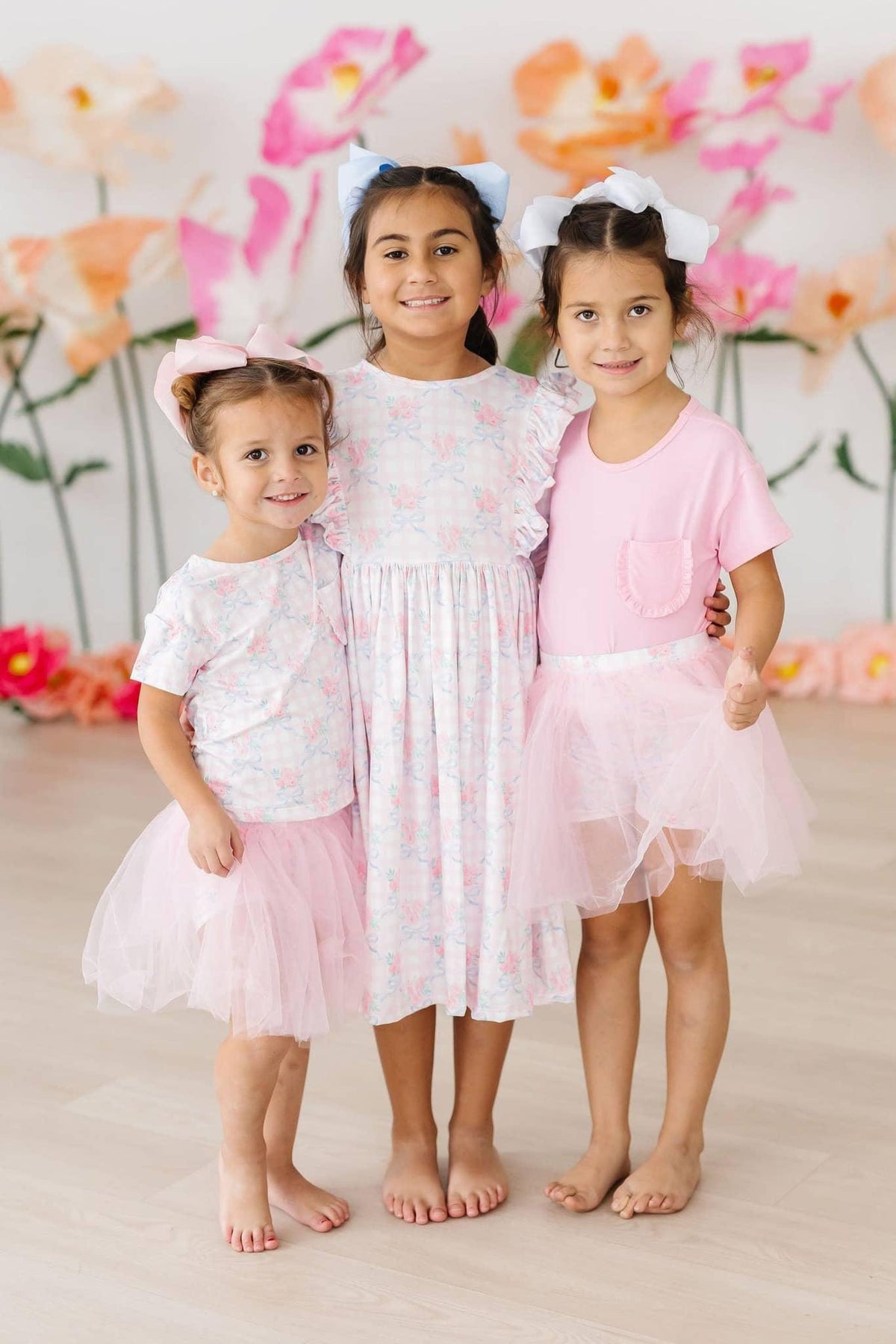 Three girls wearing girls floral ruffle pocket tee and tutus smiling and standing indoors with floral backdrop