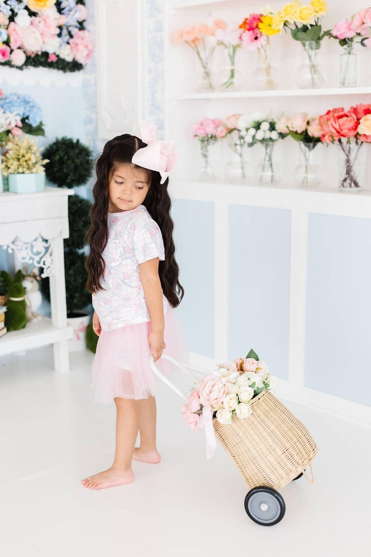 young girl wearing girls floral ruffle pocket tee and pink tulle skirt pulling a flower basket cart indoors