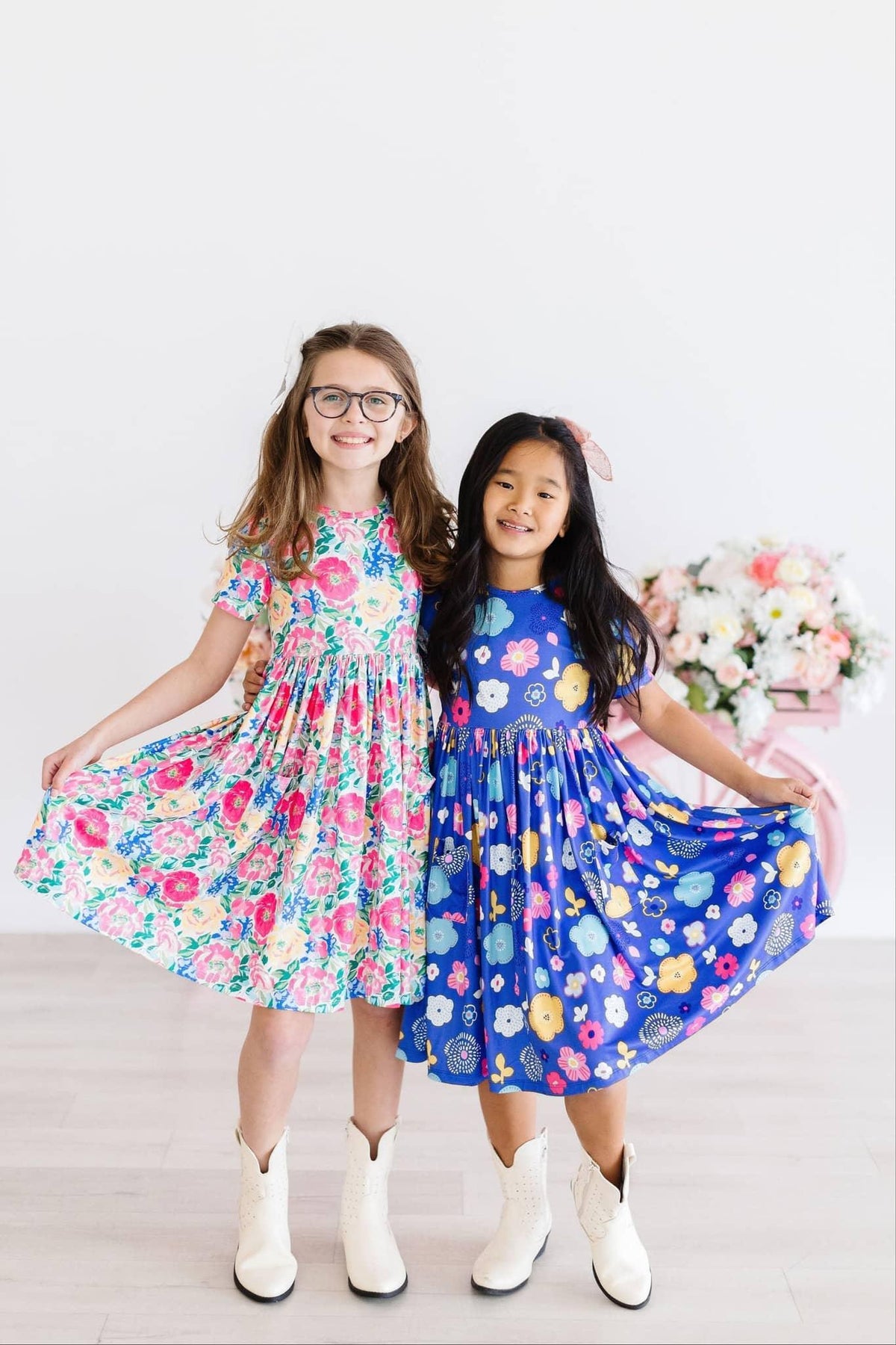 Two girls wearing colorful girls floral twirl dresses holding skirts out and smiling indoors