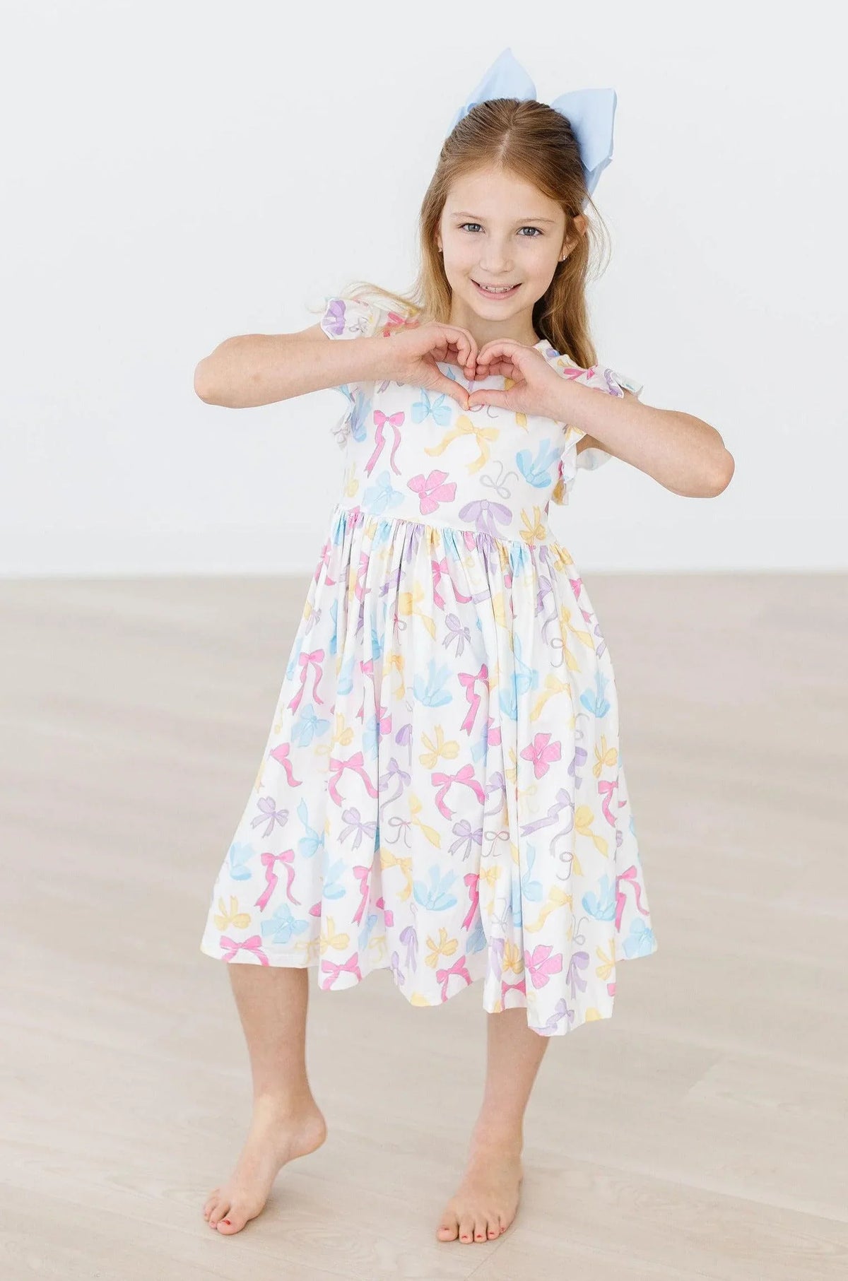 Young girl wearing a white girls flutter sleeve twirl dress with pastel bow patterns and short flutter sleeves