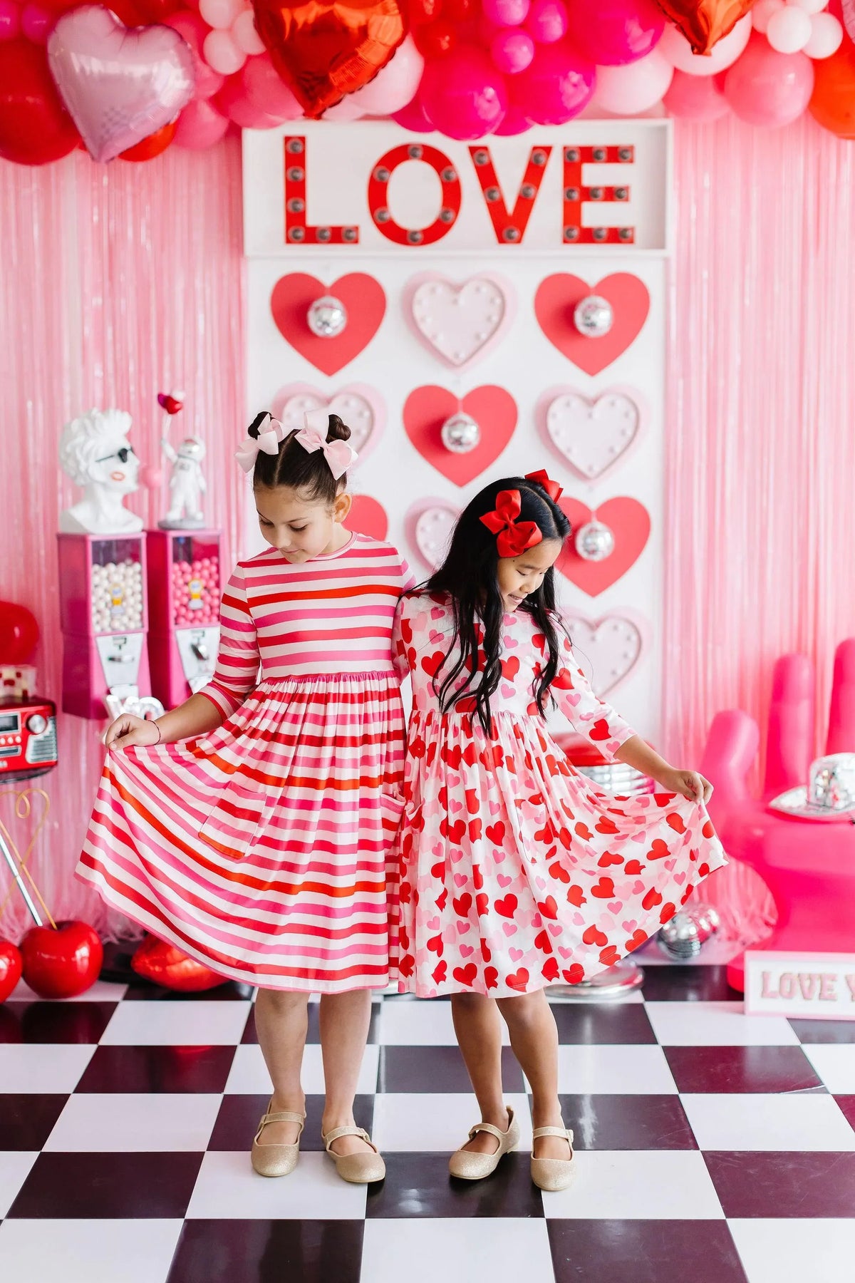 Two girls wearing girls heart print twirl dress in red and white, standing on black and white checkered floor holding dress hems