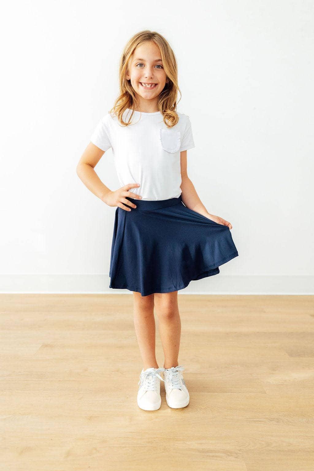 Young girl wearing girls navy skort with shorts smiling and holding the hem of the twirl skort while standing indoors
