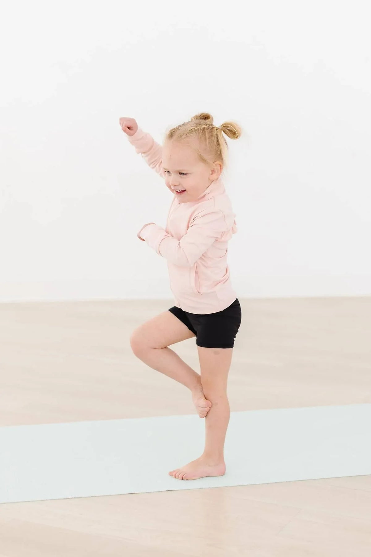 young girl wearing girls pink ruffle zip jacket and black shorts balancing on one leg indoors
