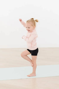 young girl wearing girls pink ruffle zip jacket and black shorts balancing on one leg indoors
