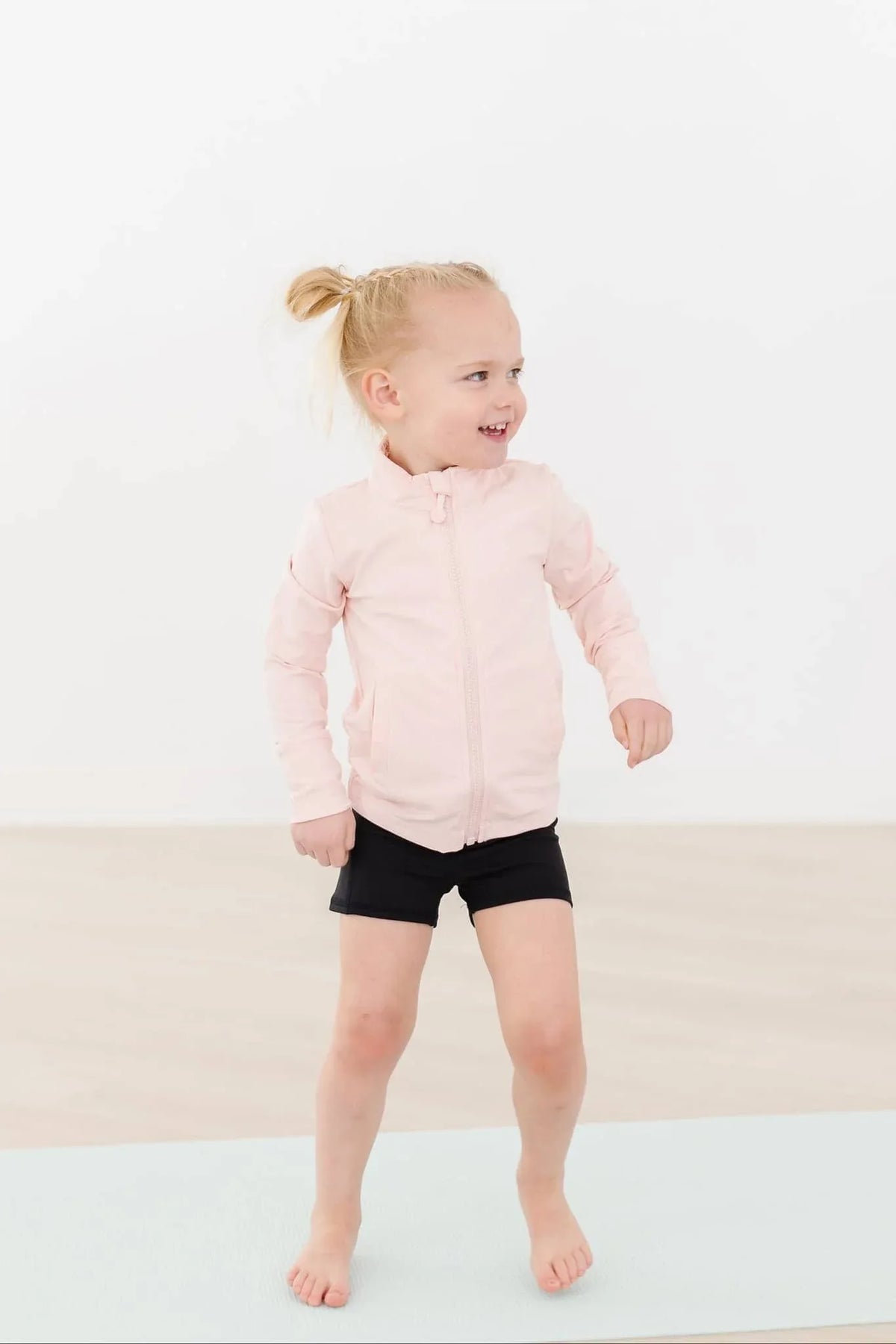 young girl wearing a girls pink ruffle zip jacket and black shorts standing and smiling indoors