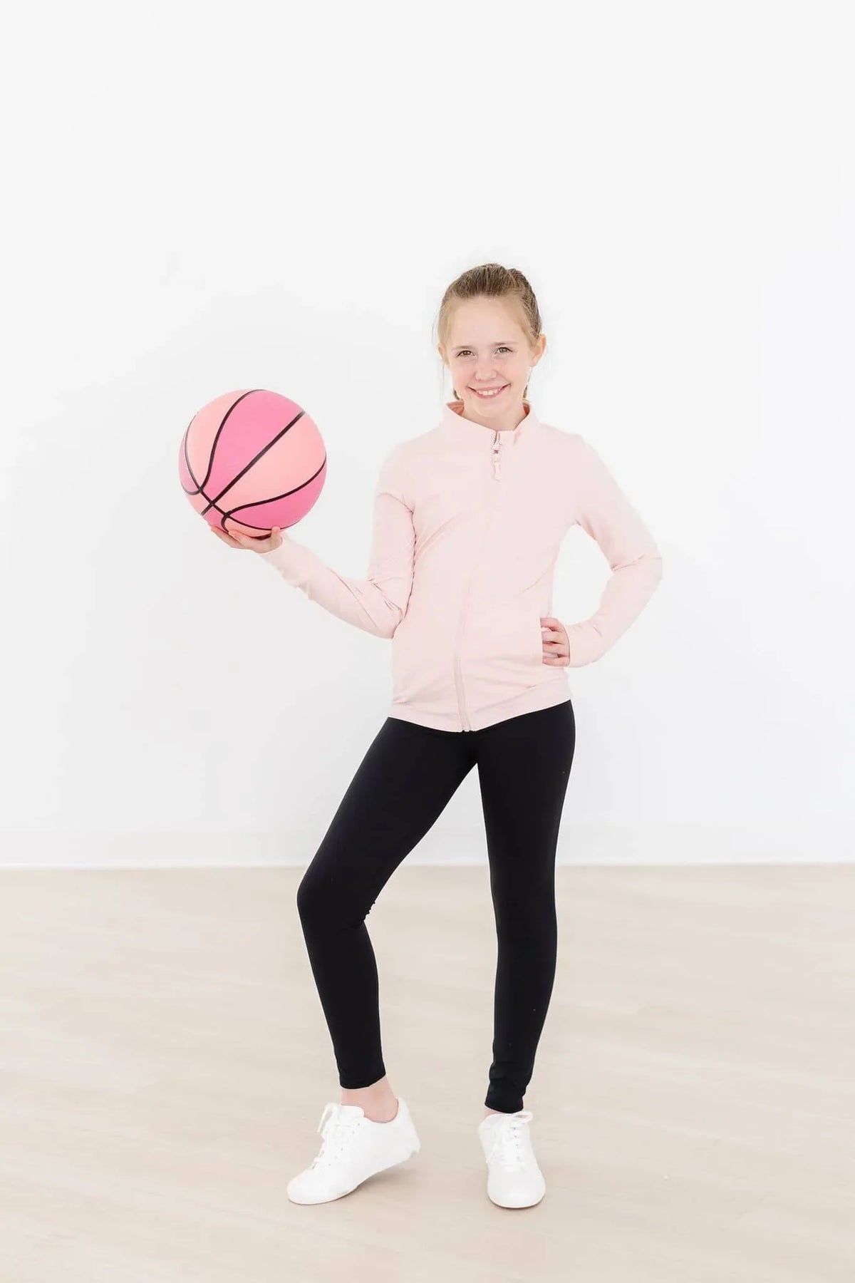 Young girl wearing girls pink ruffle zip jacket holding a pink basketball and smiling