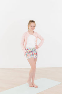 Young girl wearing girls pink ruffle zip jacket and colorful shorts standing on yoga mat smiling