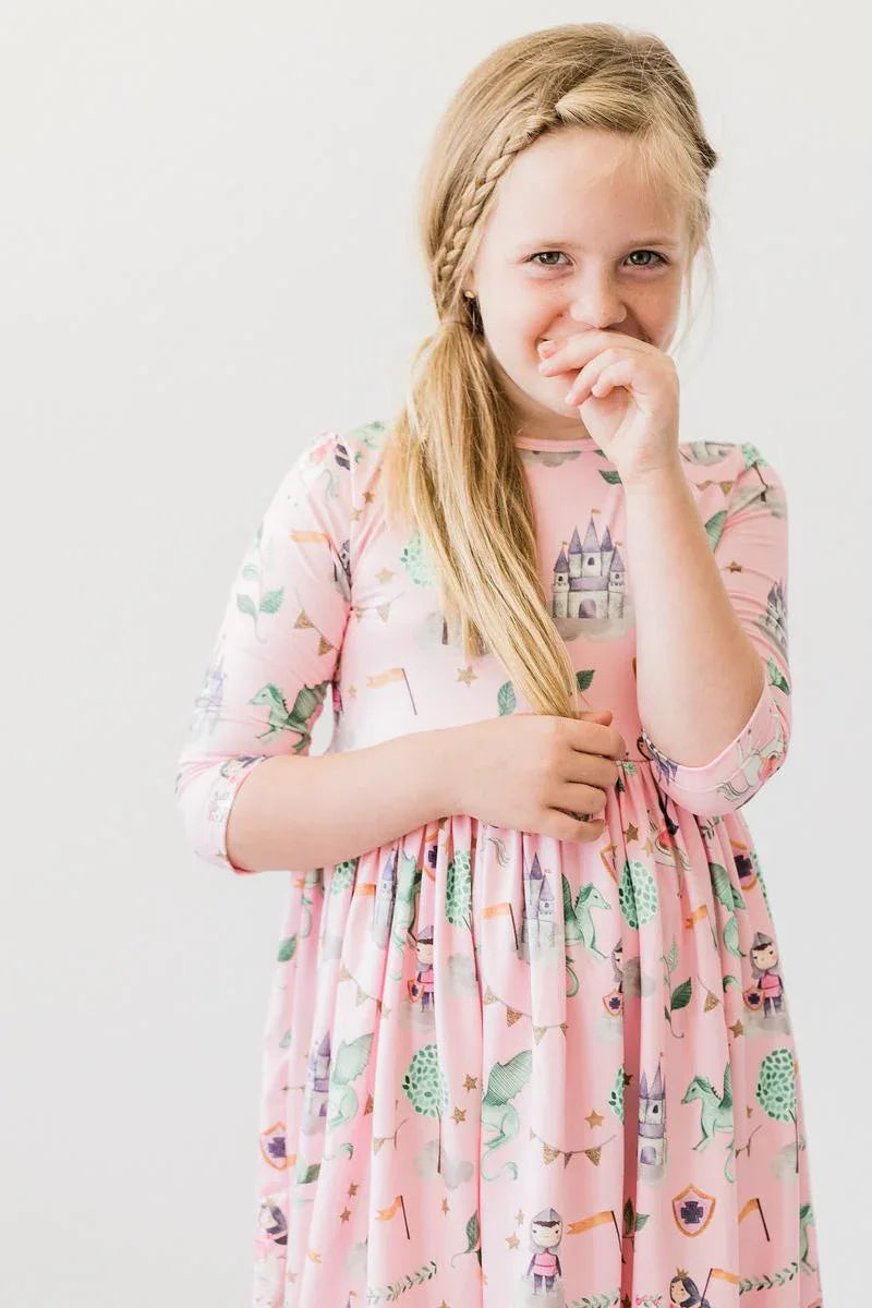 Young girl wearing pink girls pocket twirl dress with castle and unicorn prints  smiling and touching her face