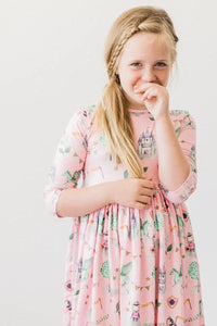 Young girl wearing pink girls pocket twirl dress with castle and unicorn prints  smiling and touching her face