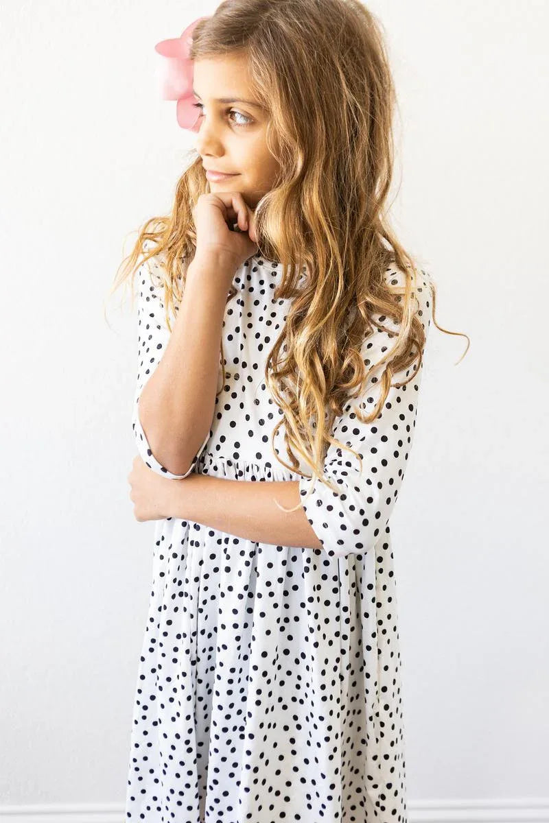 Girl wearing white and black girls polka dot twirl dress with 3/4 sleeves standing with hand near chin