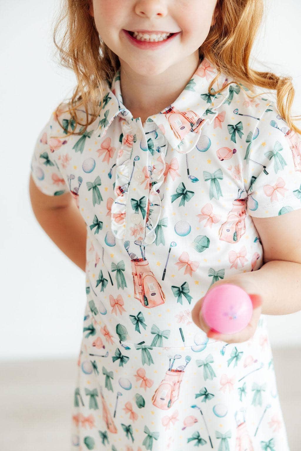 Young girl wearing a white girls polo dress with pink and green floral patterns holding a pink ball