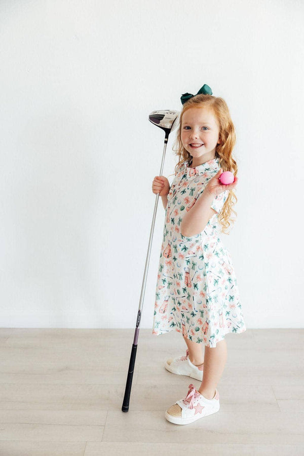 Young girl wearing white floral girls polo dress holding golf club and smiling indoors