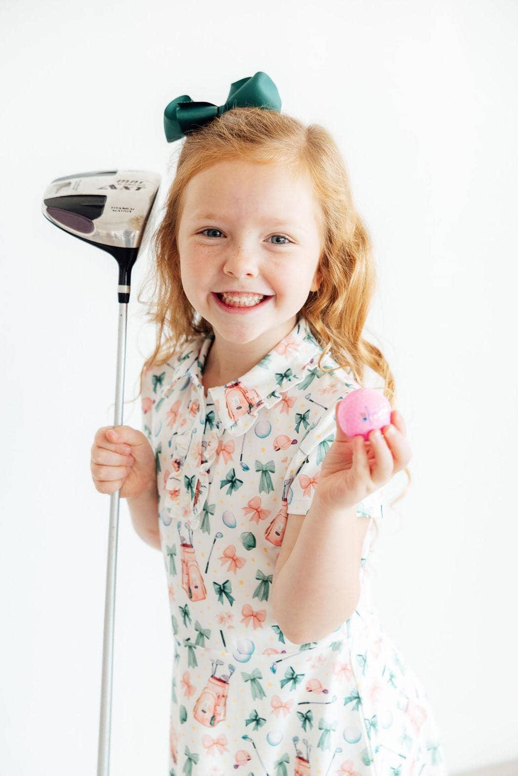 Young girl wearing a white and pastel floral girls polo dress holding a golf club and pink golf ball smiling