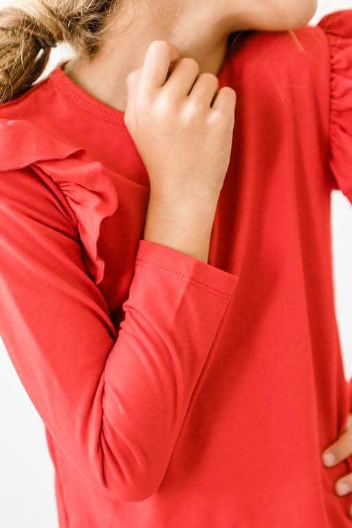 Girl wearing girls red ruffle long sleeve top with ruffle detail on shoulder posing with hand near face
