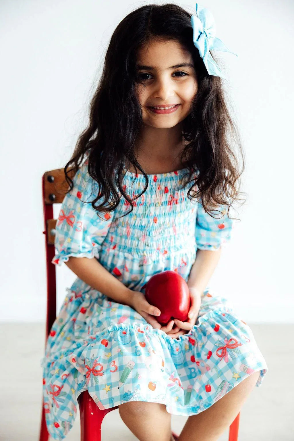 Girls smocked ruffle dress by Mila & Rose in light blue with bow print, worn by young girl with matching hair bow