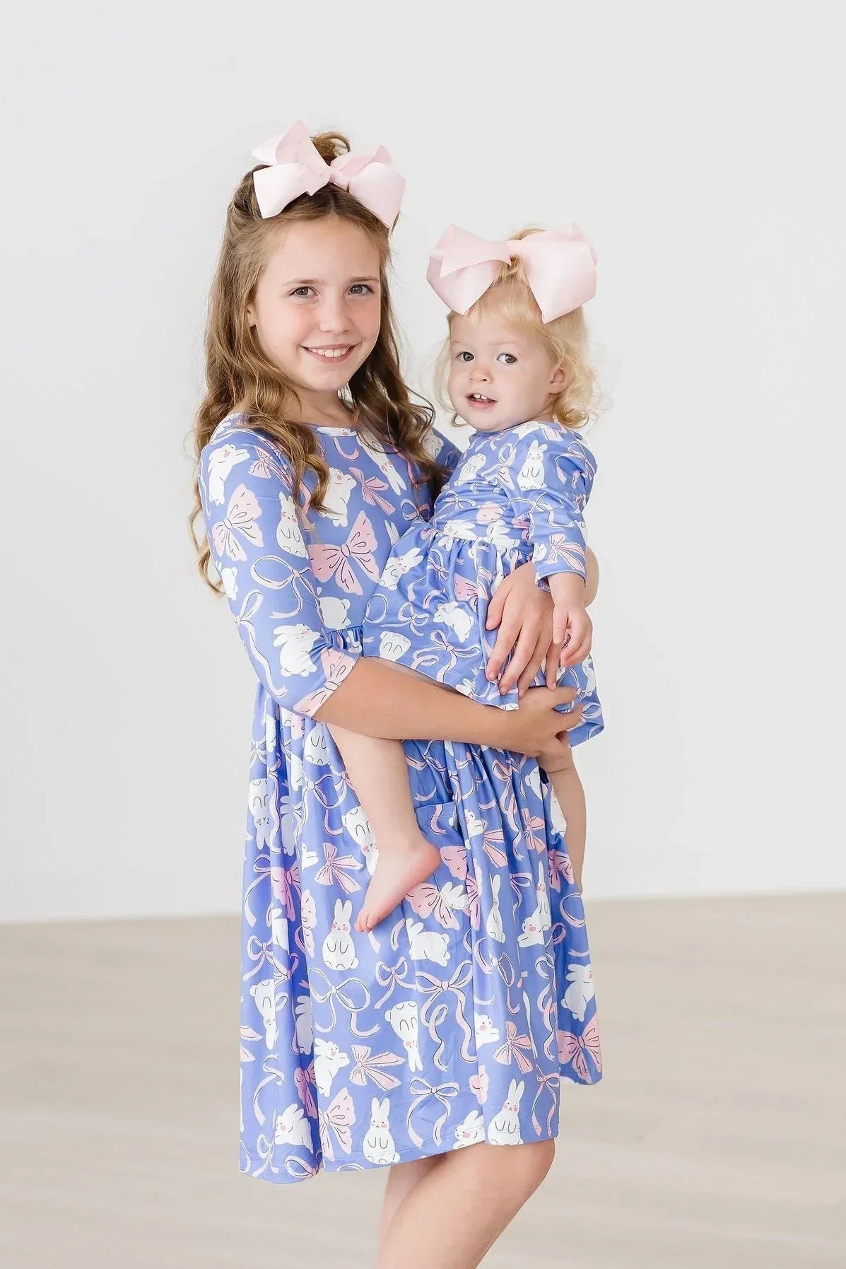 Older girl wearing a blue Mila & Rose girls twirl dress with pockets holding a toddler in a matching blue girls twirl dress with pockets