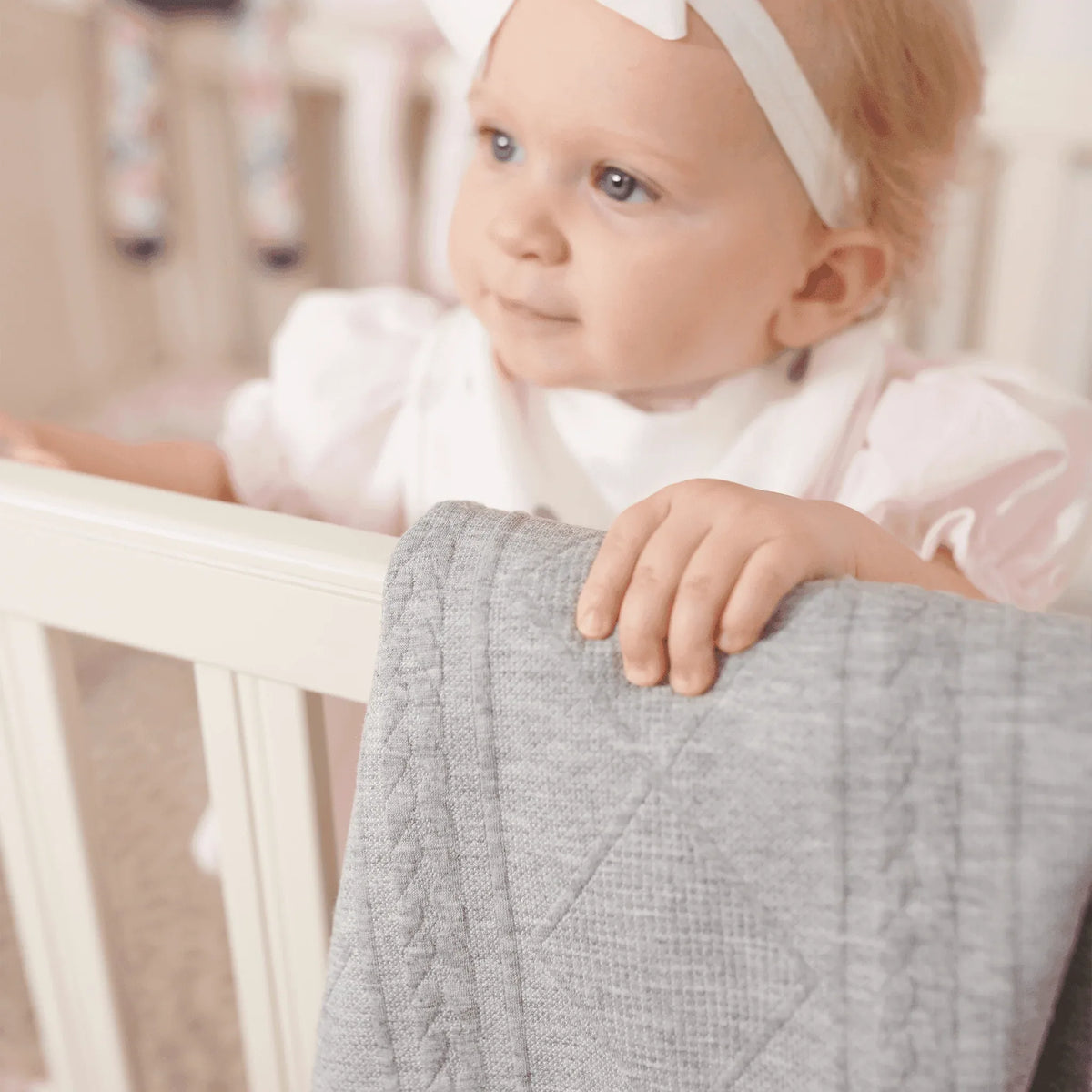 baby girl holding soft gray knit baby blanket while standing in white crib