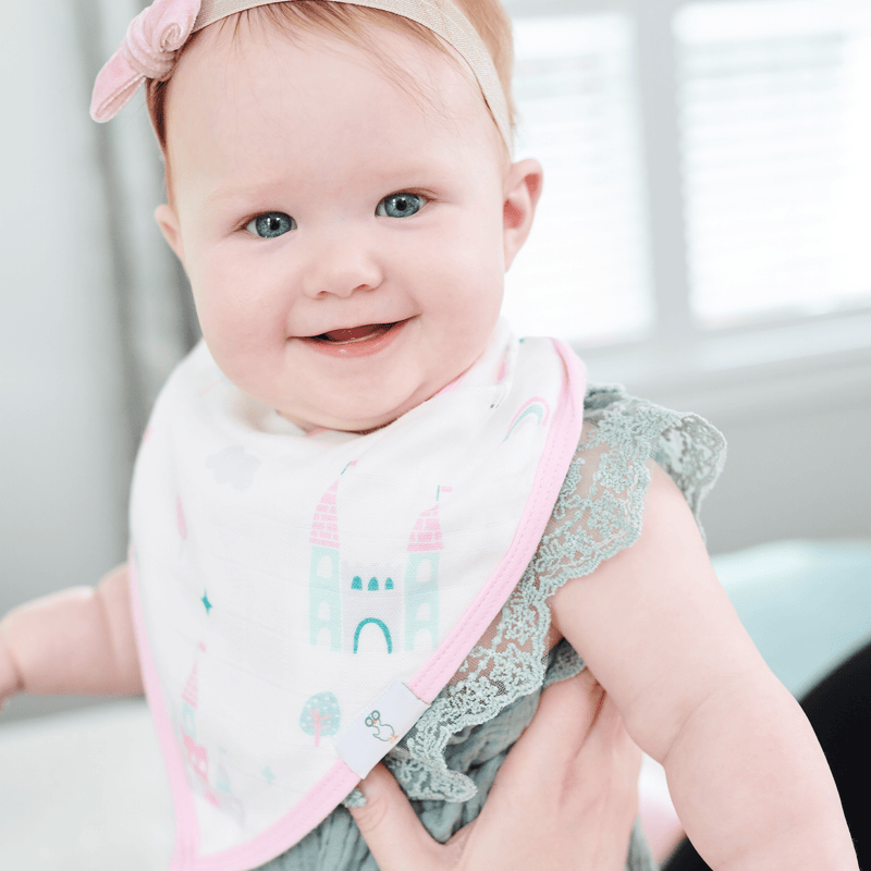 Baby wearing the Clouds and Castles 2 Pack Muslin & Terry Cloth Bib Set, smiling while showcasing the adorable bib adorned with castles and unicorns.