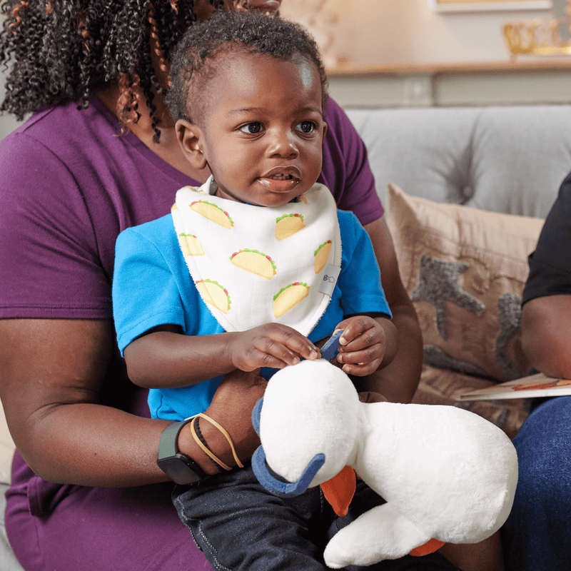 Toddler enjoying time with a soft toy while wearing the Taco Tuesday and Taco 2 Pack Muslin & Terry Cloth Bib Set, highlighting its adorable taco pattern.