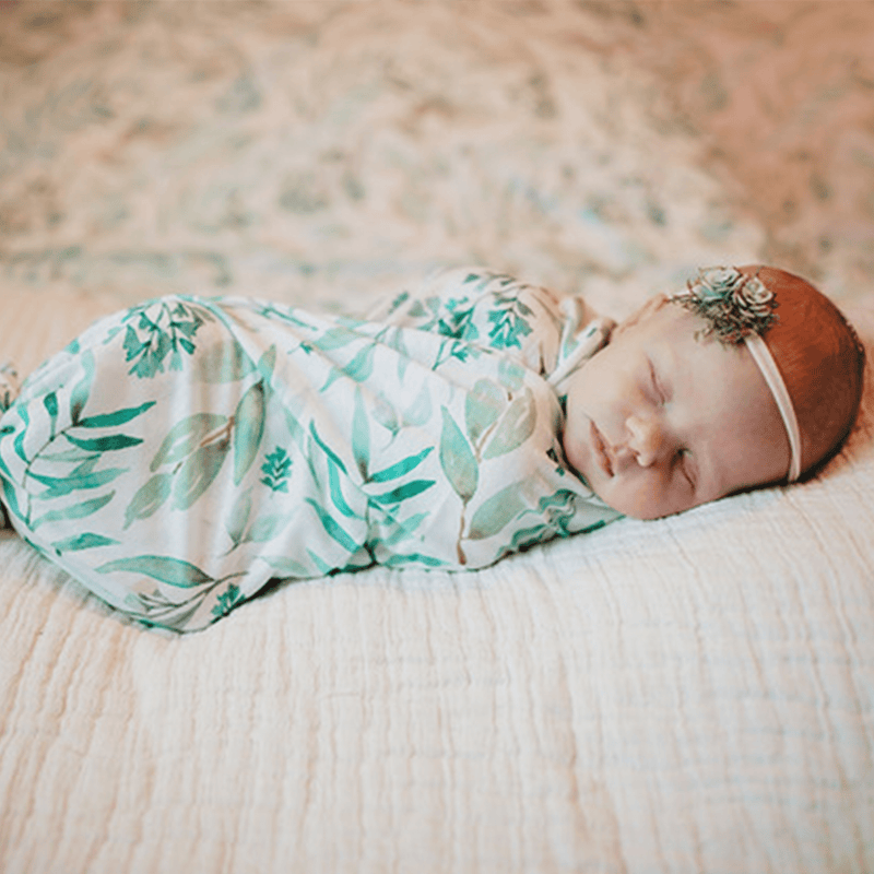 2 PK Newborn Receiving Blankets by Goosewaddle® showcasing a sweet sleeping baby wrapped in a leafy patterned swaddle, ideal for newborn comfort.