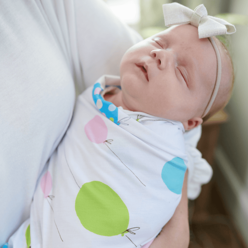 Cozy 2 Pack Newborn Receiving Blankets by Goosewaddle® featuring a swaddled baby girl sleeping peacefully, wrapped in a playful balloon print blanket. Perfect for snug comfort and versatile use.