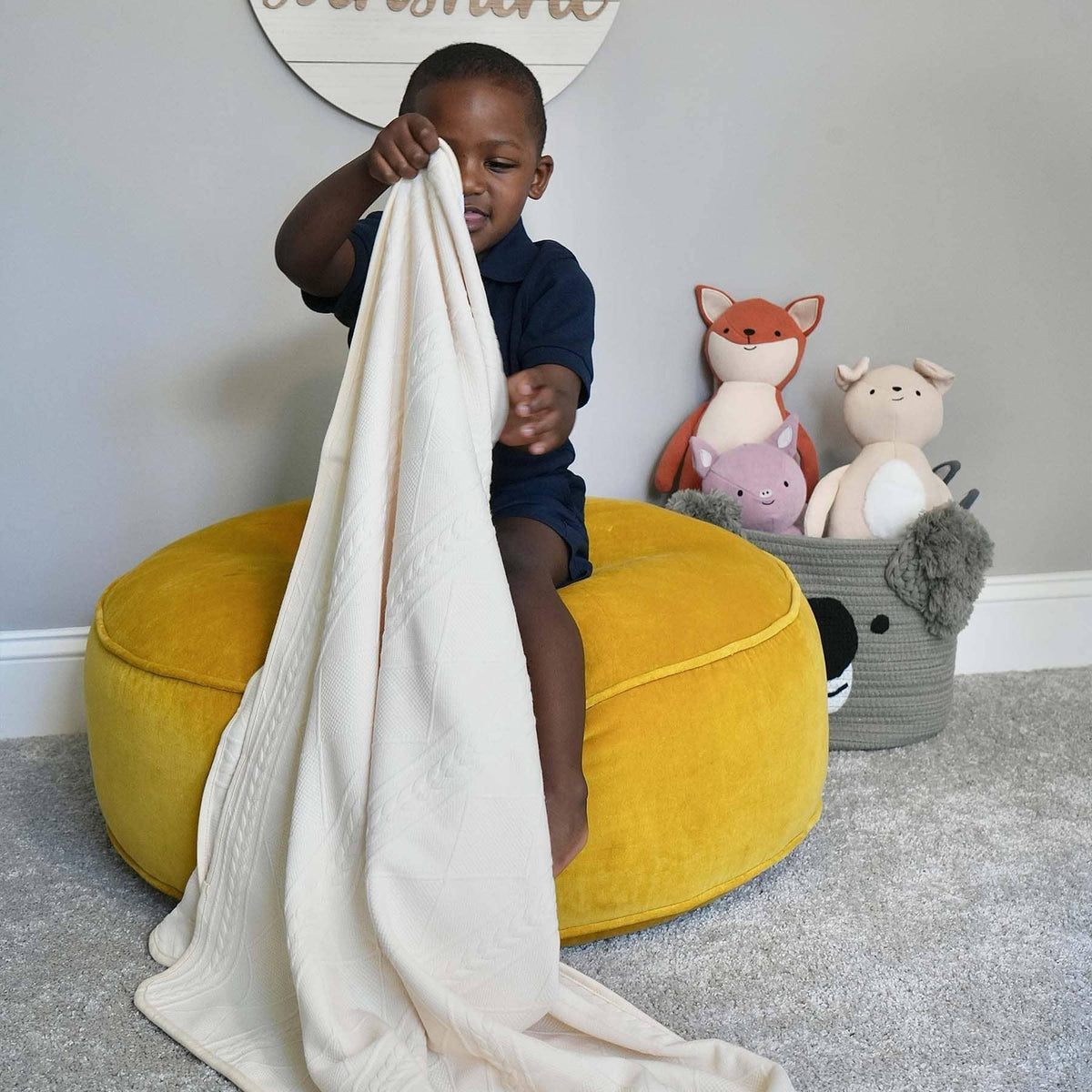toddler sitting on yellow pouf holding oatmeal knit baby blanket in a playroom